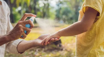 An adult sprays insect repellent on the arm of a child in an outdoor setting